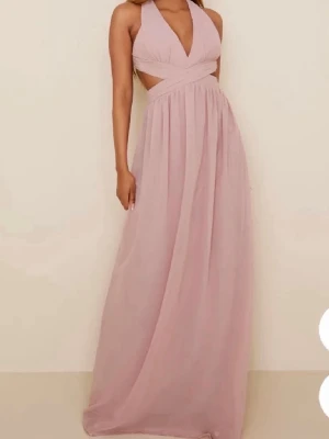 Ljusrosa långklänning med omlott - Elegant ljusrosa långklänning med omlottdetalj och öppen rygg. Klänningen har breda axelband som korsas i ryggen och en v-ringad urringning framtill. Tillverkad i ett lätt, flytande material som ger ett romantiskt intryck. Klänningen är bara använd 1 gång! 