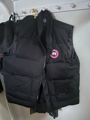 Canada Goose - Säljer en svart dunväst från Canada Goose med klassisk logotyp på bröstet. Västen har hög krage, två stora fickor framtill och dragkedja. Perfekt för kalla dagar när du vill hålla värmen och ändå se stilren ut.