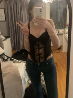 Svart spetsbody med snörning - Snygg svart body med spetsdetaljer och snörning framtill. Djup urringning och breda axelband ger en trendig look. Perfekt att styla med jeans eller kjol för en cool outfit.