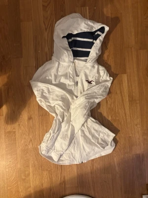 hollister tröja - Vit hoodie från Hollister med dragkedja framtill och marinblå-vita ränder i huvan. Klassisk Hollister-logga i vinrött på bröstet. Hoodien har långa ärmar och är tillverkad i mjukt bomullsmaterial. Perfekt för en avslappnad och stilren look.