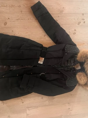 Svart Moncler kappa med pälskrage (mink) - Snygg svart kappa från Moncler med quiltade detaljer och stor pälskantad huva. Kappan har bälte i midjan, dragkedja och knappar framtill samt vadderad insida för extra värme. Nypris 16500kr