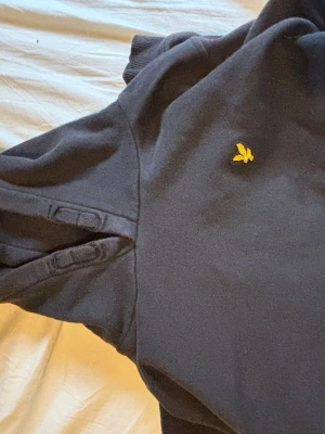 Svart sweatshirt från Lyle & Scott - Klassisk svart sweatshirt från Lyle & Scott med den ikoniska gula örnloggan broderad på bröstet. Tröjan har rund halsringning och ribbade muddar vid ärmslut och nederkant. Perfekt för en avslappnad och stilren look.