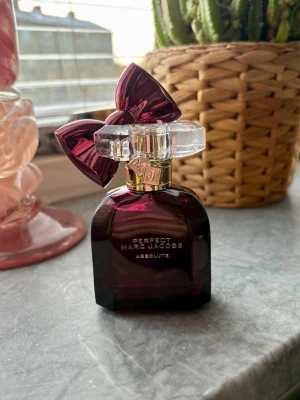 Perfect Marc Jacobs Absoluté parfym - En lyxig parfym från Marc Jacobs, Perfect Absoluté, i en mörklila glasflaska med en stor dekorativ rosett i metalliclila på korken. Flaskan har gulddetaljer och en unik, kristalliknande kork som ger en exklusiv känsla. Ny 30 ml