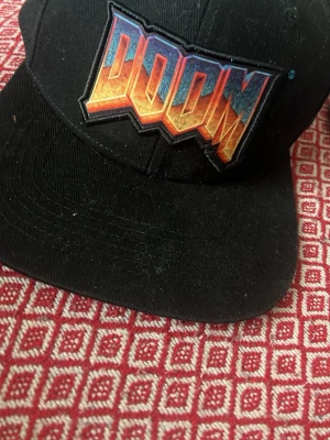Svart DOOM keps med brodyr - Säljer en svart keps med ett stort broderat DOOM-logo framtill i färgerna orange, gul och blå. Klassisk rak skärm och justerbar baksida. Perfekt för dig som gillar gaming och vill ha en unik accessoar till din stil.