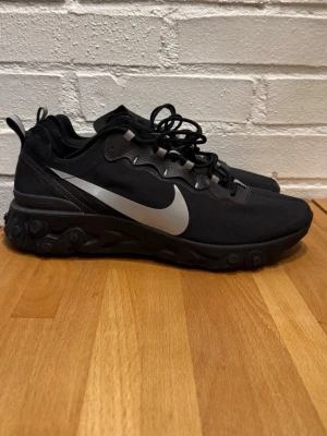 Nike React Element 55 svarta sneakers - Svarta Nike React Element 55 sneakers med vit swoosh på sidan. Skorna har en chunky sula med unika mönster och reflekterande detaljer. Ovandelen är i textil och syntet, med svarta skosnören och dragflik bak. Perfekta för dig som gillar sportig och clean stil.