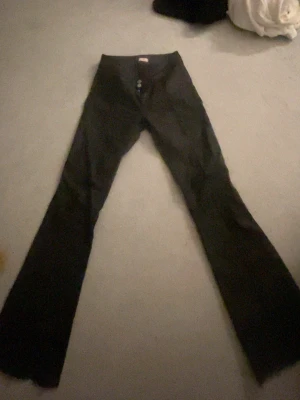 Svarta skinnbyxor Nelly 38 - Säljer ett par svarta bootcut-byxor från Nelly med två knappar och dragkedja framtill. Byxorna är ganska små i storleken jag har 36 i vanliga Nelly jeans men dessa har jag 38som sitter snyggt. Perfekta för dig som gillar en trendig siluett.