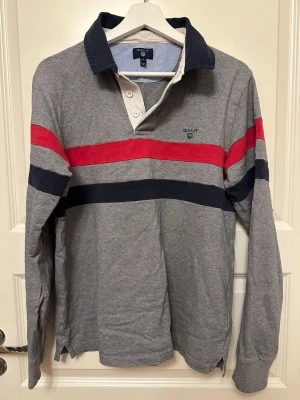 Rugbytröja från Gant - Långärmad rugbytröja från Gant i grått med breda röda och marinblå ränder över bröstet och ärmarna. Tröjan har en mörkblå krage, knappar vid halsen och är gjord i mjuk bomull. Perfekt för en sportig och avslappnad look.