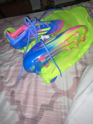 Adidas blåa och rosa fotbollsskor - Säljer ett par Adidas fotbollsskor i blått, rosa och neongult med klassiska tre ränder på sidan. Skorna har snörning, låg profil och dobbar för spel på gräs. Materialet är syntetiskt och designen är riktigt sportig och modern. Perfekta för dig som vill sticka ut på planen.