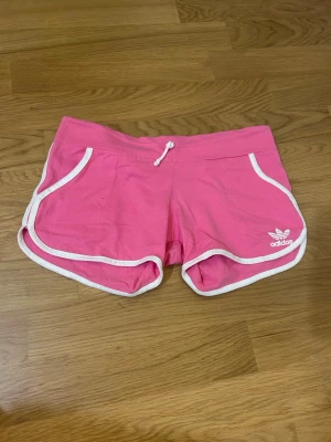 Rosa Adidas shorts med vita detaljer - Snygga rosa shorts från Adidas med vita kanter och snörning i midjan. Klassisk Adidas-logga framtill och stor logga baktill. Perfekta för träning eller chill. Tillverkade i mjuk bomull för skön känsla. De är lite längre ner där bak