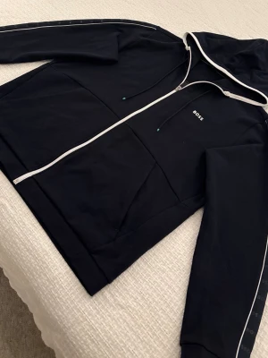 Mörkblå Hugo Boss zip hoodie - Zip hoodie från Hugo Boss i mjuk bomullsblandning. Modell med hel dragkedja, huva och sidofickor på vardera sida. Diskret BOSS-logotyp på bröstet samt logoband med upprepade BOSS-tryck längs ärmarna.  Färg: mörkblå (navy). Storlek XL (INT). Mycket bra skick, inga fläckar eller hål.  Äkta BOSS – inneretiketter och detaljer finns med på bilderna.