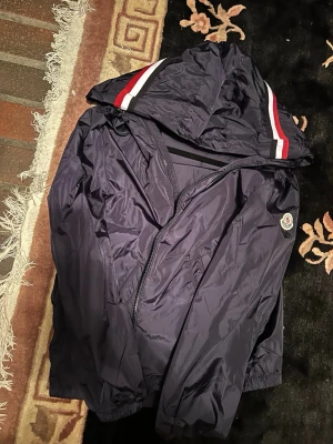 Mörkblå Moncler vindjacka med huva - Snygg mörkblå vindjacka från Moncler med huva och dragkedja. Jackan har vita och röda ränder på huvan samt Moncler-logga på ärmen. Tillverkad i lätt och glansigt material som står emot vind. Perfekt för dig som vill ha en stilren och sportig look.