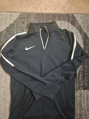 Svart Nike träningströja med dragkedja - Snygg svart långärmad träningströja från Nike med vit logga på bröstet och vita ränder längs ärmarna. Tröjan har hög krage och halv dragkedja framtill. Perfekt för träning eller uppvärmning. Tillverkad i ett mjukt och stretchigt funktionsmaterial.