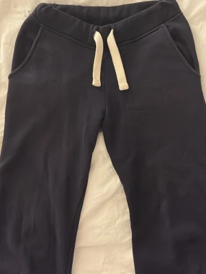 Mörkblå mjukisbyxor från russemerch - Säljer ett par mörkblå mjukisbyxor med vita snören i midjan och två sidofickor. Byxorna har en avslappnad passform och är gjorda i mjukt bomullsmaterial, perfekta för chill dagar. Klassisk design utan synligt märke. ena sida av snöret är igensytt!! 