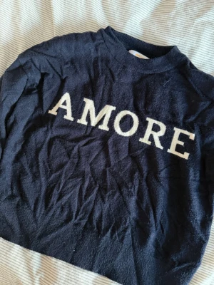 Mörkblå tröja med AMORE-tryck - Mysig mörkblå tröja med vit text 'AMORE' på bröstet. Tröjan har rund halsringning och långa ärmar. Perfekt för dig som gillar enkel men snygg stil.