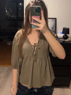 Grön blus med snörning - Säljer en grön blus med trekvartsärm och snygg snörning framtill. Blusen har en lös passform och ett lätt, luftigt tyg som ger en avslappnad vibe. Perfekt att styla med jeans eller kjol för en trendig look.