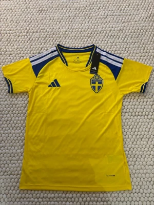 Svenska landslagets fotbollströja Adidas - Gul fotbollströja från Adidas med blå detaljer och Sveriges landslagsemblem på bröstet. Tröjan har klassiska Adidas-ränder på axlarna och är tillverkad i lätt funktionsmaterial. Perfekt för träning eller match.