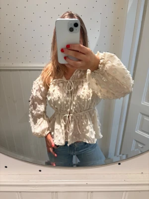 Vit blus med struktur från Lindex - Supersöt vit blus från Lindex med struktur och broderade detaljer. Blusen har långa puffärmar, knytband i midjan och vid halsen samt en lätt volang nedtill. Tillverkad i ett tunt, luftigt material som känns riktigt fräscht och trendigt.