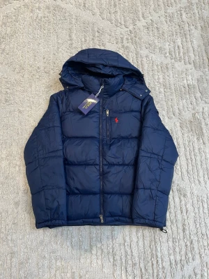 Mörkblå pufferjacka från Polo Ralph Lauren - Snygg mörkblå pufferjacka från Polo Ralph Lauren med huva och dragkedja framtill. Jackan har en liten röd logga på bröstet och quiltad design. Perfekt för kalla dagar och riktigt skön att bära. Materialet är glansigt och ger en modern look.