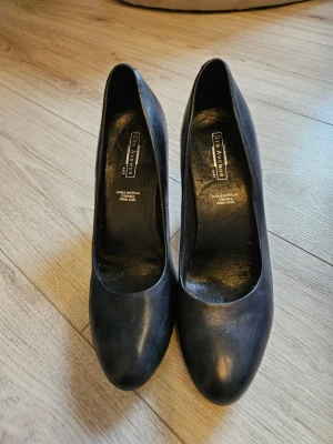 Svarta pumps från 5th Avenue - Klassiska svarta pumps från 5th Avenue i äkta läder. Skorna har rundad tå och en elegant hög klack som ger en stilren siluett. Perfekta för att lyfta vilken outfit som helst med en tidlös look.