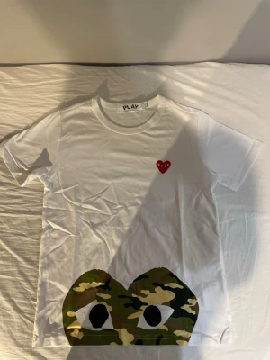 Vit Comme des Garçons PLAY t-shirt - Vit t-shirt från Comme des Garçons PLAY med det ikoniska röda hjärtat på bröstet och ett stort camo-tryck med ögon längst ner. Klassisk rund hals och korta ärmar. Perfekt statement-plagg för dig som gillar streetwear och unika prints.