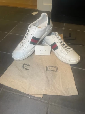 Gucci ace  - Säljer ett par klassiska vita sneakers från Gucci med den ikoniska röd-gröna randen på sidan och grönt parti vid hälen. Skorna är i läder och har vita snören samt vit sula. Medföljer originalpåse och kort. Perfekta för dig som vill ha en stilren och lyxig look. Pris kan diskuteras, vertifikat finns och vid frågor är det bara att fråga