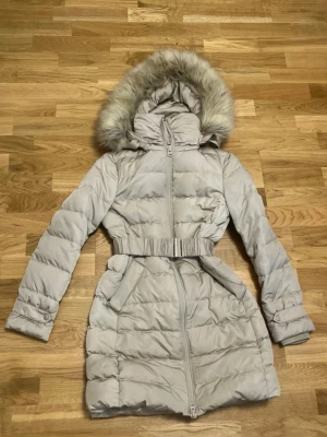 Beige dunjacka med fuskpäls från Zara - Lång, beige dunjacka från Zara med avtagbar huva och fuskpäls. Jackan har bälte i midjan, två fickor framtill och dragkedja. Perfekt för kalla vinterdagar och har en stilren, puffig design.