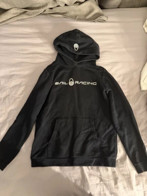 Svart Sail Racing hoodie med tryck - Svart hoodie från Sail Racing med vit logga tryckt på bröstet och en mindre logga på huvan. Klassisk känguruficka framtill och ribbade muddar vid ärmslut och nederkant. Tillverkad i mjukt bomullsmaterial för skön känsla.