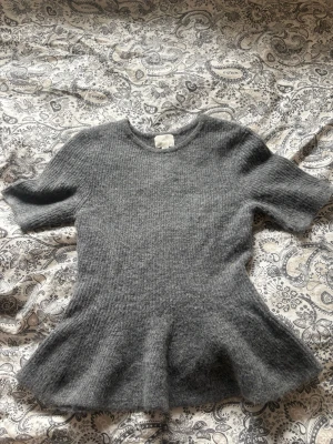 Grå stickad topp från H&M - Mysig grå stickad topp från H&M med korta ärmar och rund hals. Modellen har en lätt utsvängd midja som ger en snygg siluett. Använd vid endast två tillfällen.
