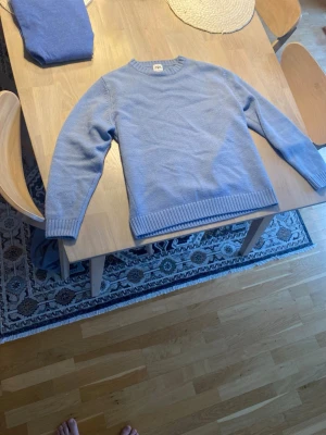 Ljusblå stickad tröja från Arket - Säljer en ljusblå stickad tröja från Arket med rund halsringning och ribbade muddar vid ärmslut och nederkant. Tröjan har en clean och enkel design som passar perfekt till jeans eller kjol. Materialet känns mjukt och bekvämt.