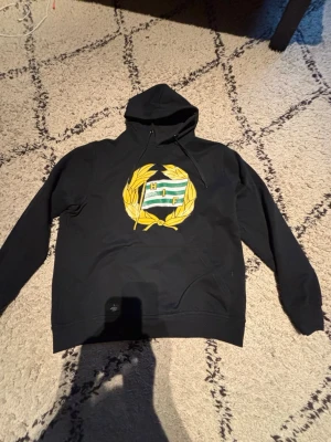 Svart Hammarby hoodie med emblem - Svart hoodie från Hammarby med stort broderat klubbemblem i grönt, vitt och gult på bröstet. Klassisk huva med dragsko och känguruficka framtill. Tillverkad i mjukt bomullsmaterial, perfekt för dig som vill visa ditt stöd för Bajen.