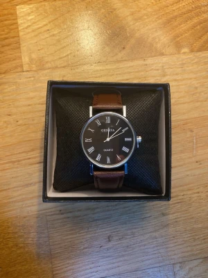 Klassisk klocka från Geneva brun/svart - Stilren klocka från Geneva med svart urtavla, romerska siffror och silverfärgade visare. Armbandet är brunt och ser ut att vara i skinn. Klockan har en rund form och klassisk design som passar till många stilar.