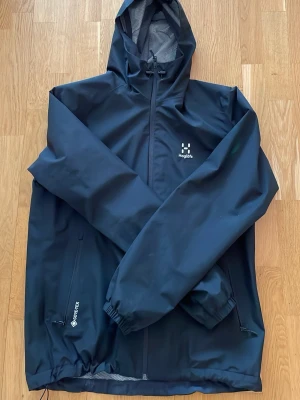 Svart regnjacka från Haglöfs - Haglöfsjacka i vattentätt material. Den har stora fickor och är justerbar i midja och luva