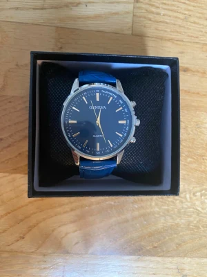 Blå klocka från Geneva med silverdetaljer - Stilren klocka från Geneva med marinblå urtavla och matchande blått armband. Klockan har silverfärgade detaljer och klassisk rund form. Armbandet är i syntetmaterial med struktur och urtavlan har tydliga markeringar för timmar och minuter.