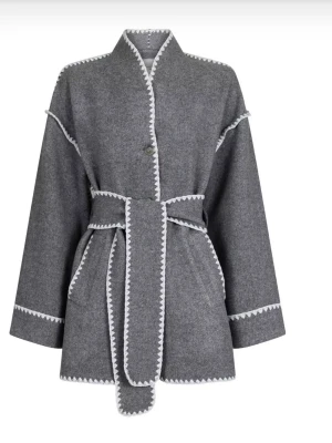 Grå ullkappa med vit kant från neo noir - Stilren grå kappa i ullmix med markerade vita kanter och knytskärp i midjan. Rak passform, lång ärm och dekorativa detaljer längs sömmarna. Perfekt för dig som gillar minimalistisk och trendig look.