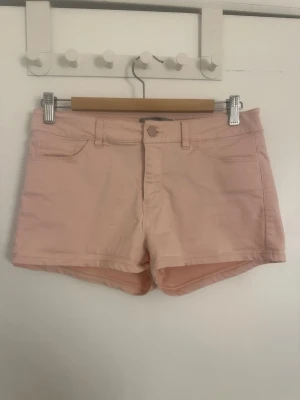 Ljusrosa jeansshorts med fickor - Säljer ett par ljusrosa shorts i jeansmodell med klassiska fickor fram och bak samt hällor för skärp. Perfekta för varma dagar och har en snygg, enkel design som är lätt att matcha. Liten defekt vid ena bakfickan (se bild)