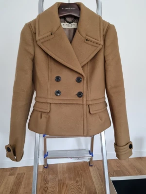 Klassisk beige skepparkavaj Burberry - Snygg beige skepparkavaj från Burberry med dubbelknäppning och bred krage. Superexklusiv känsla och material. Jackan har dekorativa knappar vid ärmslut och markerad midja. Tillverkad i ull för en lyxig känsla och tidlös stil. Perfekt för dig som vill ha en ikonisk jacka i garderoben.