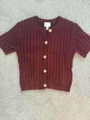 Vinröd kabelstickad kofta H&M - Kortärmad vinröd kabelstickad kofta från H&M med guldfärgade knappar framtill. Tröjan har rund halsringning och ribbade kanter. Perfekt för lager på lager och ger en trendig touch till din outfit.