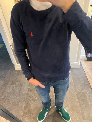 Mörkblå tröja  från Ralph Lauren - Mörkblå sweatshirt från Ralph Lauren med klassisk rund halsringning och den ikoniska röda logotypen broderad på bröstet. Tröjan har långa ärmar och ribbade muddar vid ärmslut och nederkant. Perfekt för en avslappnad och stilren look.