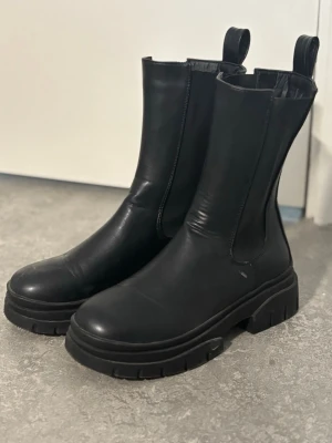 Svarta chunky boots i skinn - Säljer ett par svarta boots med chunky sula och högt skaft. Tillverkade i slätt skinn med elastiska paneler på sidorna och dragflikar upptill för enkel påtagning. Perfekta för dig som gillar en stilren men edgy look.