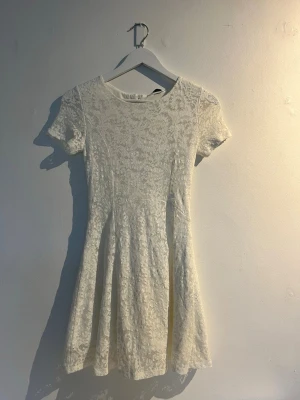 Vit spetsklänning med kort ärm - Söt vit kortklänning med spetsdetaljer och rund hals. Klänningen har korta ärmar och en lätt utställd kjol som ger en feminin siluett. Dragkedja i ryggen för enkel påklädning. Storlek 158/164 