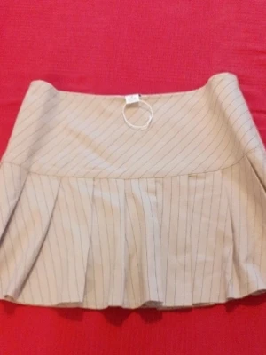 Beige kortkjol med tunna ränder - Snygg beige kortkjol med tunna svarta ränder och veckad nederdel. Kjolen har en hög midja och ett stilrent, modernt uttryck. Perfekt för dig som gillar en trendig och enkel look.