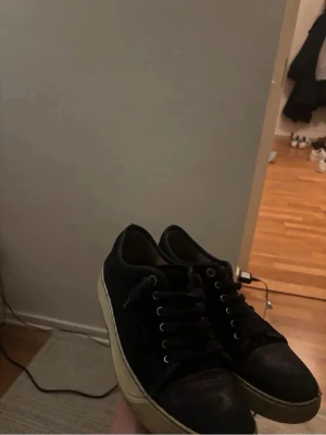 Svarta Lanvin sneakers i mocka - Säljer ett par svarta sneakers från Lanvin med ovandel i mocka och vita gummisulor. Skorna har rund tå, snörning med metallöljetter och klassisk låg profil. Perfekta för dig som gillar stilrena och tidlösa sneakers.