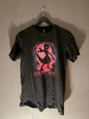 Svart Pokémon Mewtwo T-shirt - Cool svart T-shirt med stort tryck av Mewtwo från Pokémon i rosa och rött framtill. Rund halsringning och korta ärmar. Perfekt för dig som älskar gaming och anime.