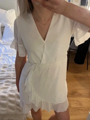Vit omlottklänning med volang - Snygg vit kort omlottklänning med volangdetaljer och v-ringning. Använd endast ett tillfälle, i mycket bra skick! Perfekt till studenten eller andra somriga tillfällen!