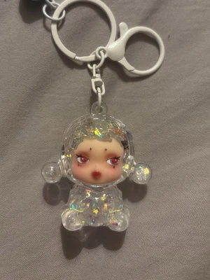 Glittrig vattenfylld skullpanda nyckelring  - Säljer en söt nyckelring med en genomskinlig figur fylld med glitter och små färgade detaljer. Nyckelringen är i plast och har en rundad form med armar och ben. Perfekt att hänga på väskan eller nycklarna för en extra touch.