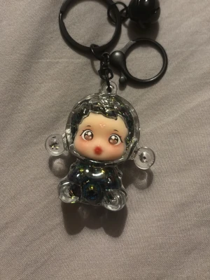 Glittrig vatten fylld skullpanda nyckelring  - Säljer en söt nyckelring med en genomskinlig figur i akryl fylld med glitter och små blommor. Nyckelringen har svarta ringar och en liten bjällra. Perfekt att hänga på väskan eller nycklarna för en unik touch.