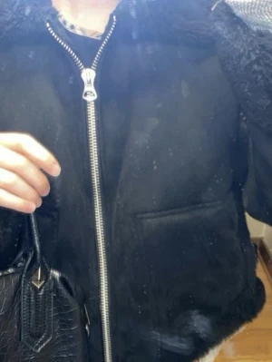 Svart teddy kappa med dragkedja - Snygg svart teddy kappa med silvrig dragkedja framtill och mjukt, fluffigt material. Jackan har en avslappnad passform och breda muddar. Perfekt för att hålla värmen under kyliga dagar och ger en trendig look.