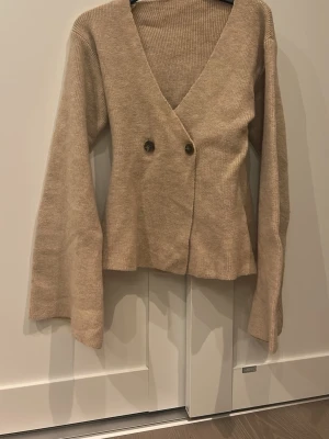 Beige stickad kofta med v-ringning - Beige stickad kofta med djup v-ringning och två knappar framtill. Koftan har långa ärmar och en avslappnad passform, perfekt för lager på lager. Enkel och stilren design som passar till det mesta.