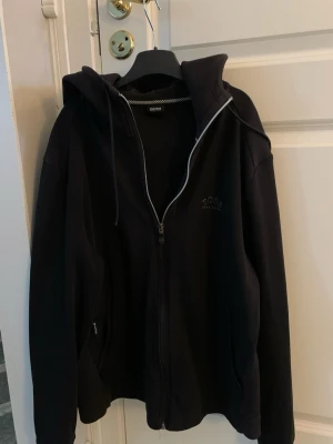 Svart Hugo boss hoodie med dragkedja från BOSS - Pris kan diskuteras vid snabbt köp👍Snygg svart hoodie från BOSS med dragkedja framtill och justerbar huva med snören. Diskret BOSS-logga broderad på bröstet. Hoodien har långa ärmar och ribbade muddar. Perfekt för en avslappnad och stilren look.