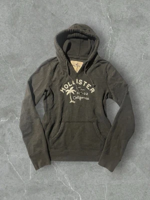 Mörkgrå hoodie från Hollister | 40/L - Mysig mörkgrå hoodie från Hollister med broderad logga, palm och texten 'California' på bröstet. Hoodien har snörning i huvan och en klassisk känguruficka framtill. Perfekt för chill dagar och enkel att matcha med jeans eller mjukis.
